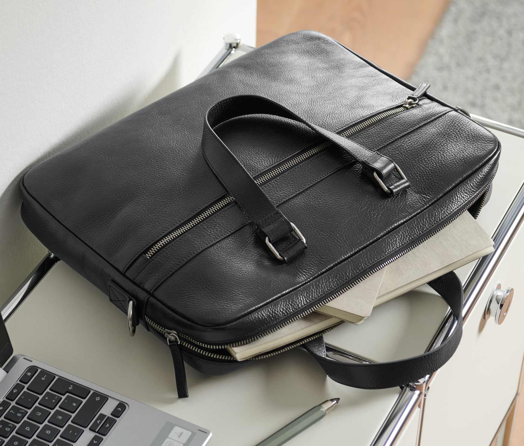 Eine schwarze Leder-Aktentasche liegt neben einem Laptop und einem Stift auf einem Tisch.