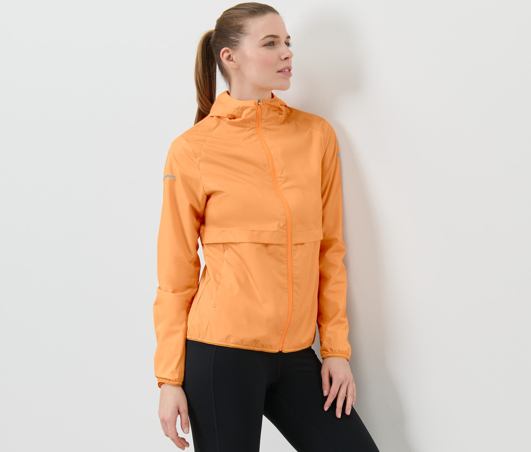 Frau posiert in neonoranger Windprotection-Laufjacke und schwarzen Leggings.