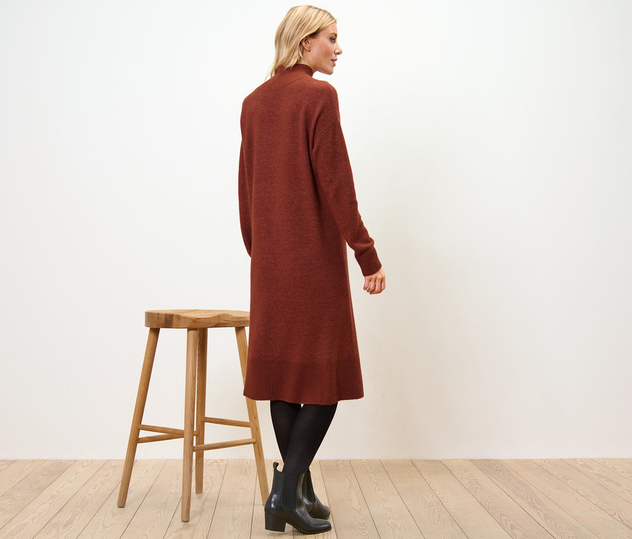 Frau steht seitlich und trägt ein Strickkleid mit Wollanteil, eine schwarze Strumpfhose und Stiefeletten. Neben ihr steht ein Holzhocker.