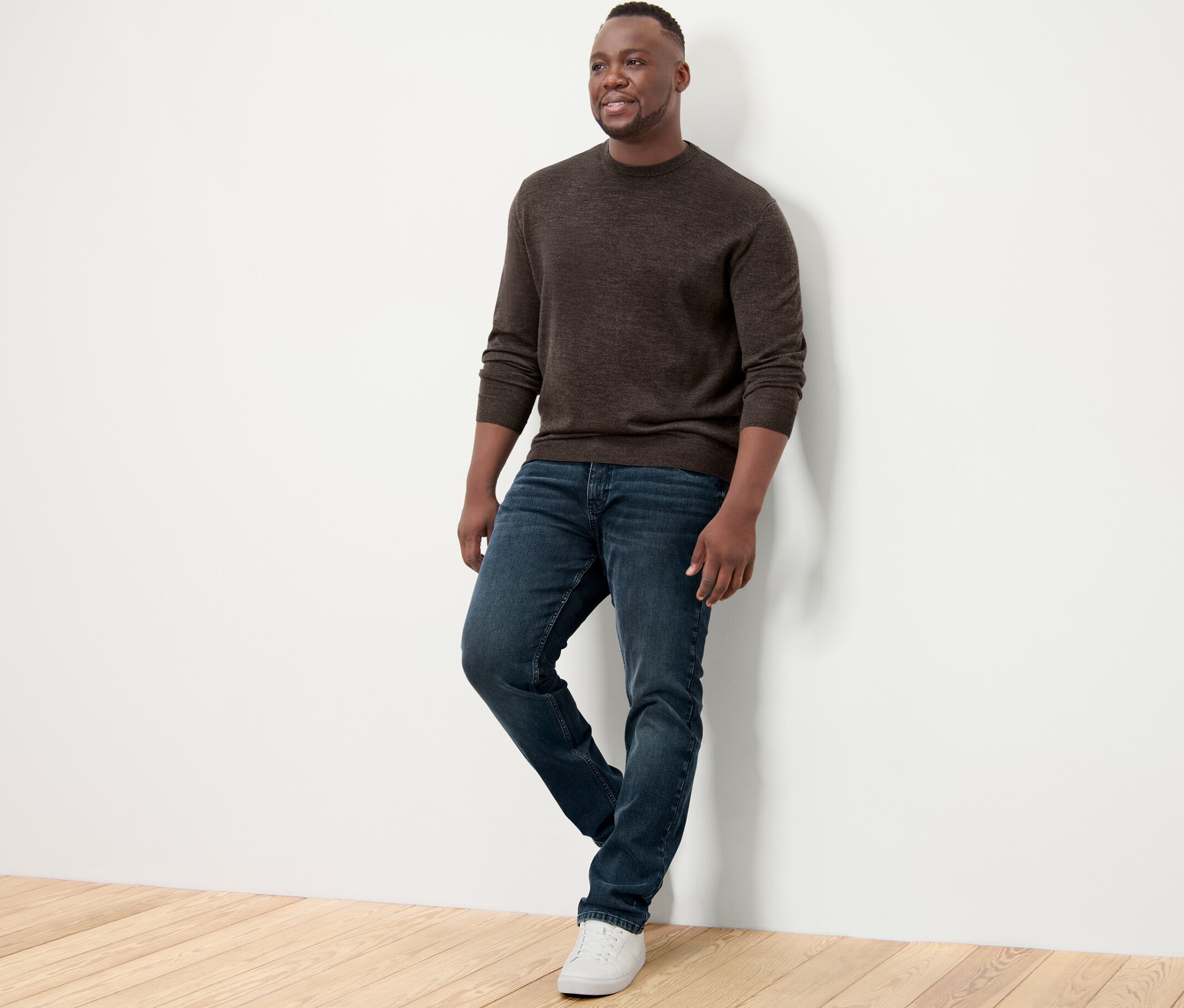 Mann an eine Wand gelehnt, trägt einen braunen Merino-Pullover, Jeans und Turnschuhe.