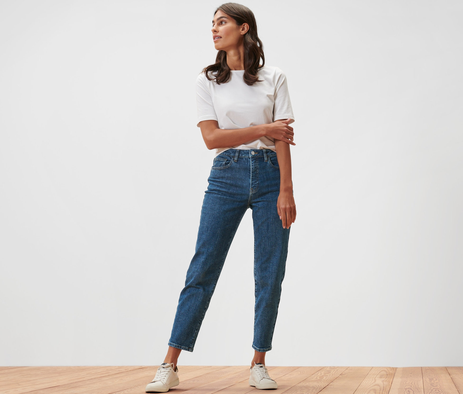 Eine Frau steht auf einem Holzboden und trägt Mom Jeans – Fit »Nyla« und ein weißes T-Shirt.