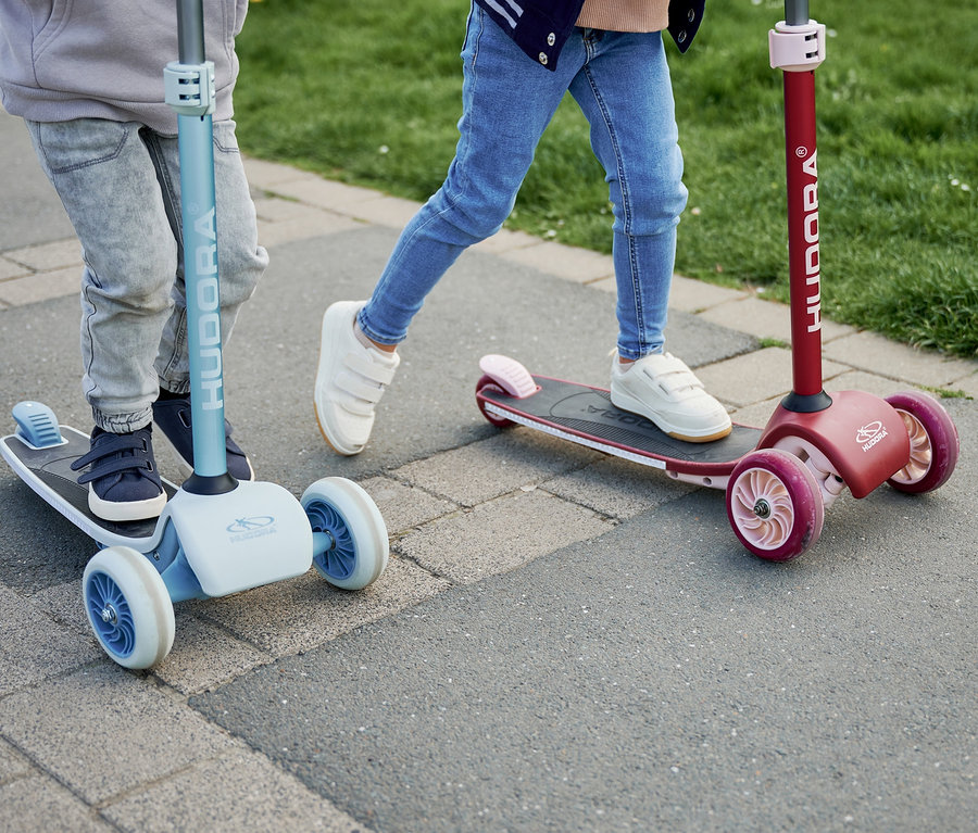 Zwei Kinder fahren mit HUDORA Tri-Scootern, einer bordeauxrot, der andere blau, auf dem Gehweg.