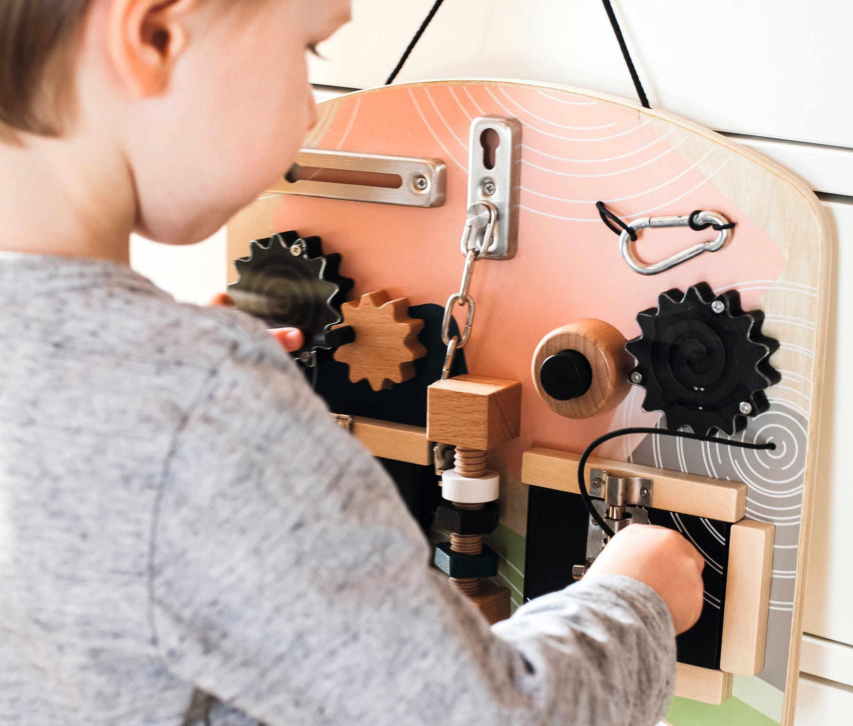 Ein Junge spielt mit dem small foot Motorikbrett »Schlösser und Rotation«, das an einem weißen Schrank befestigt ist.