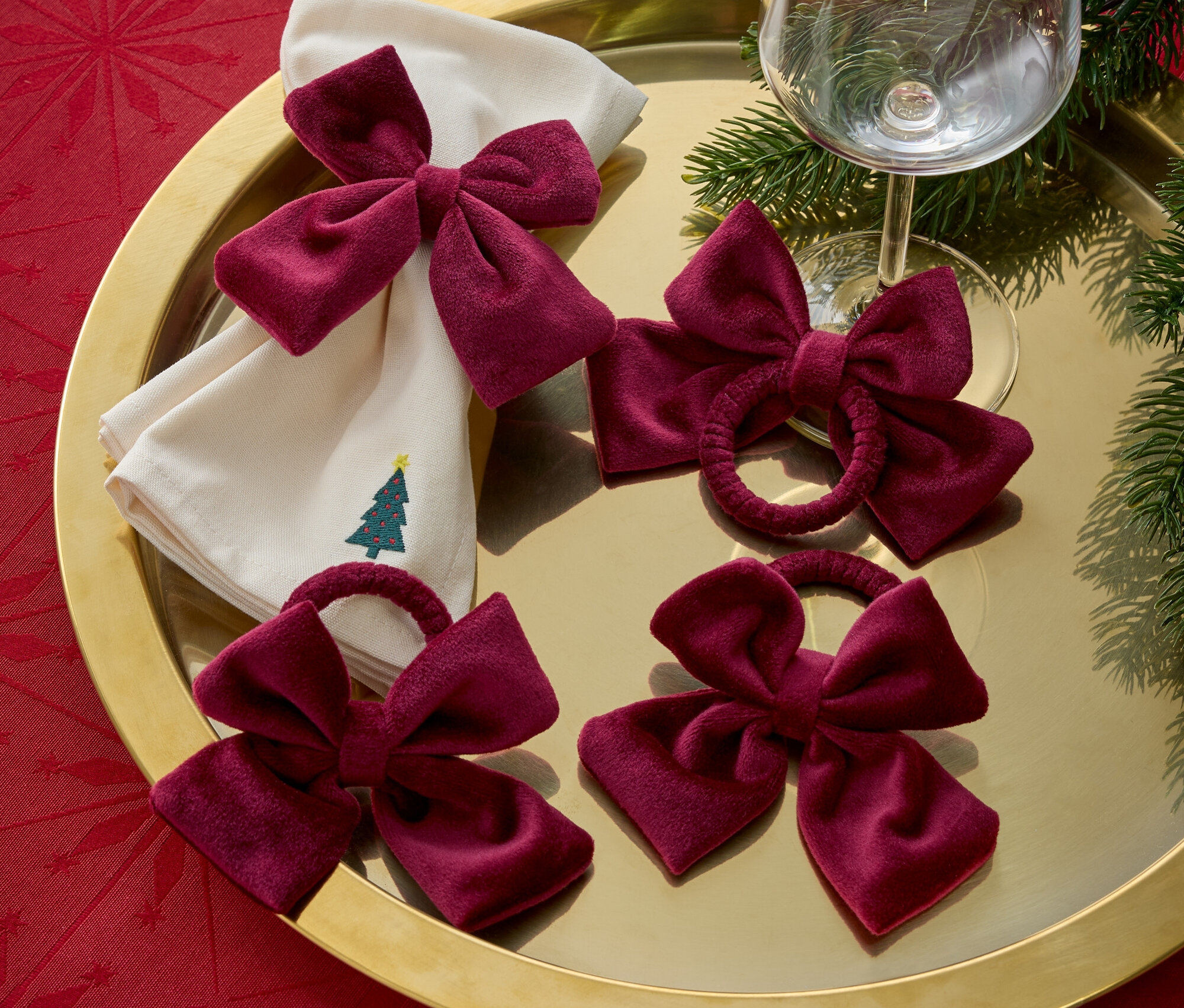 Auf einem goldenen Tablett liegen Stoffservietten mit Weihnachtsbaumstickerei, bordeauxrote Schleifen und ein Weinglas.