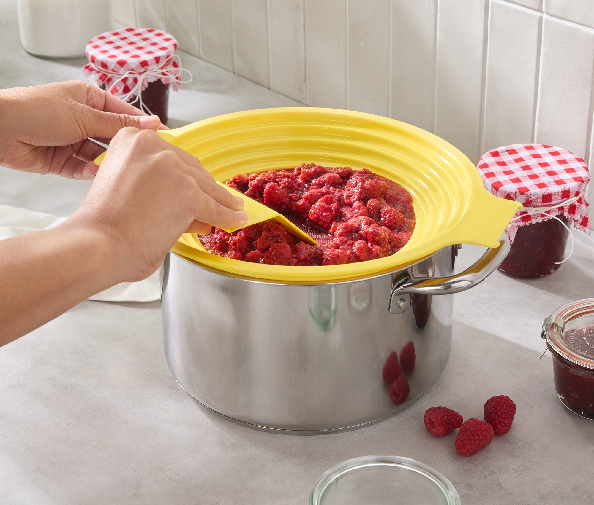 Eine Person rührt Himbeermarmelade in einem Topf mit einem gelben Spritzschutz. Einige Himbeeren und Gläser stehen auf der Küchentheke.