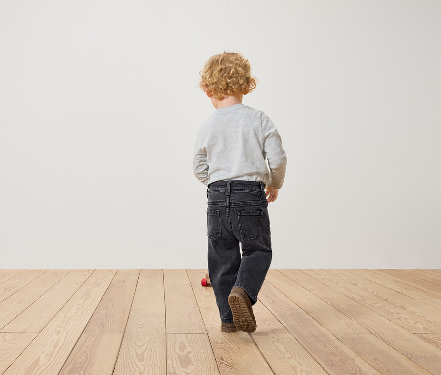 Junge mit lockigem Haar in grauem T-Shirt und schwarzen Jeans geht auf einem Holzboden.