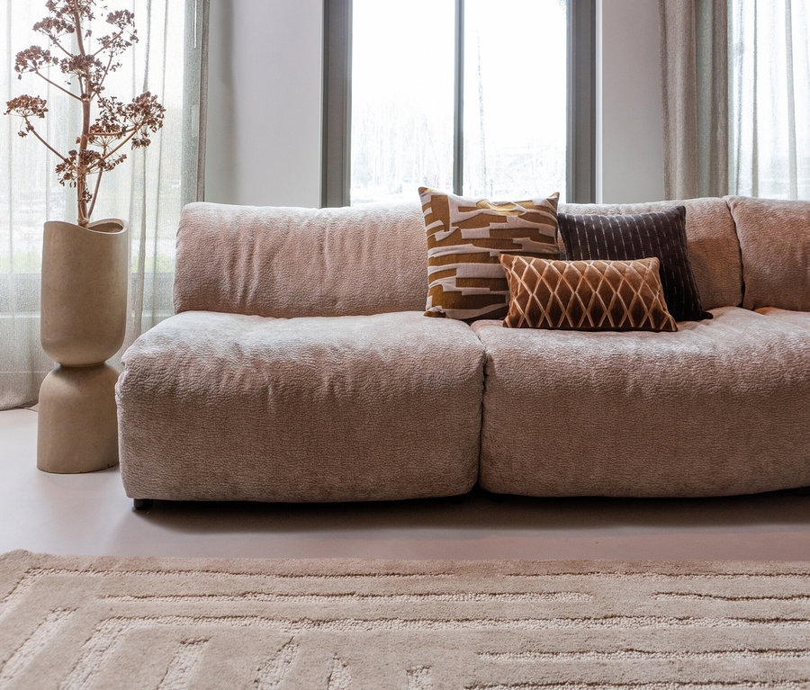 Ein beiges Sofa mit drei Kissen steht in einem Wohnzimmer mit beigen Vorhängen und einer Blume in einer Vase, sowie einem beigen Woood Alicia Teppich.