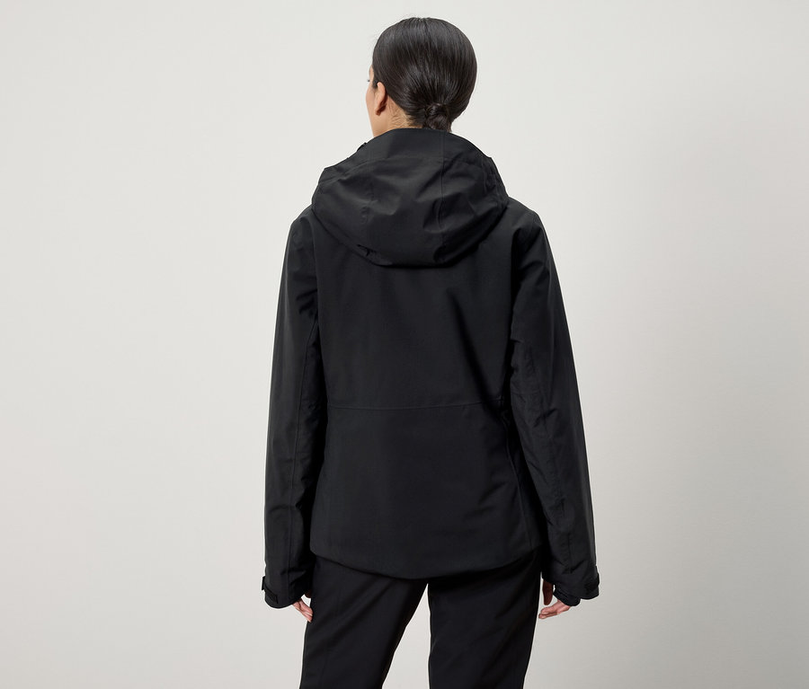 Von hinten ist eine Frau mit zum Pferdeschwanz hochgestecktem Haar zu sehen, die eine schwarze Kapuzenjacke und eine schwarze Hose trägt.