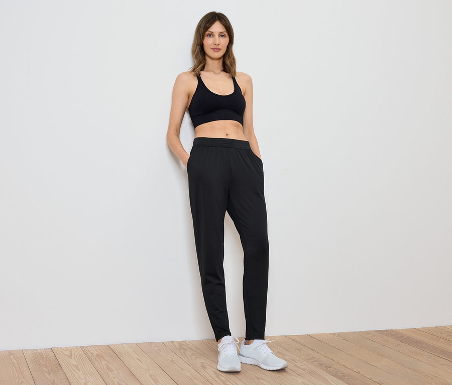 Eine Frau in einem schwarzen Sporttop und Hosen steht an einer weißen Wand.