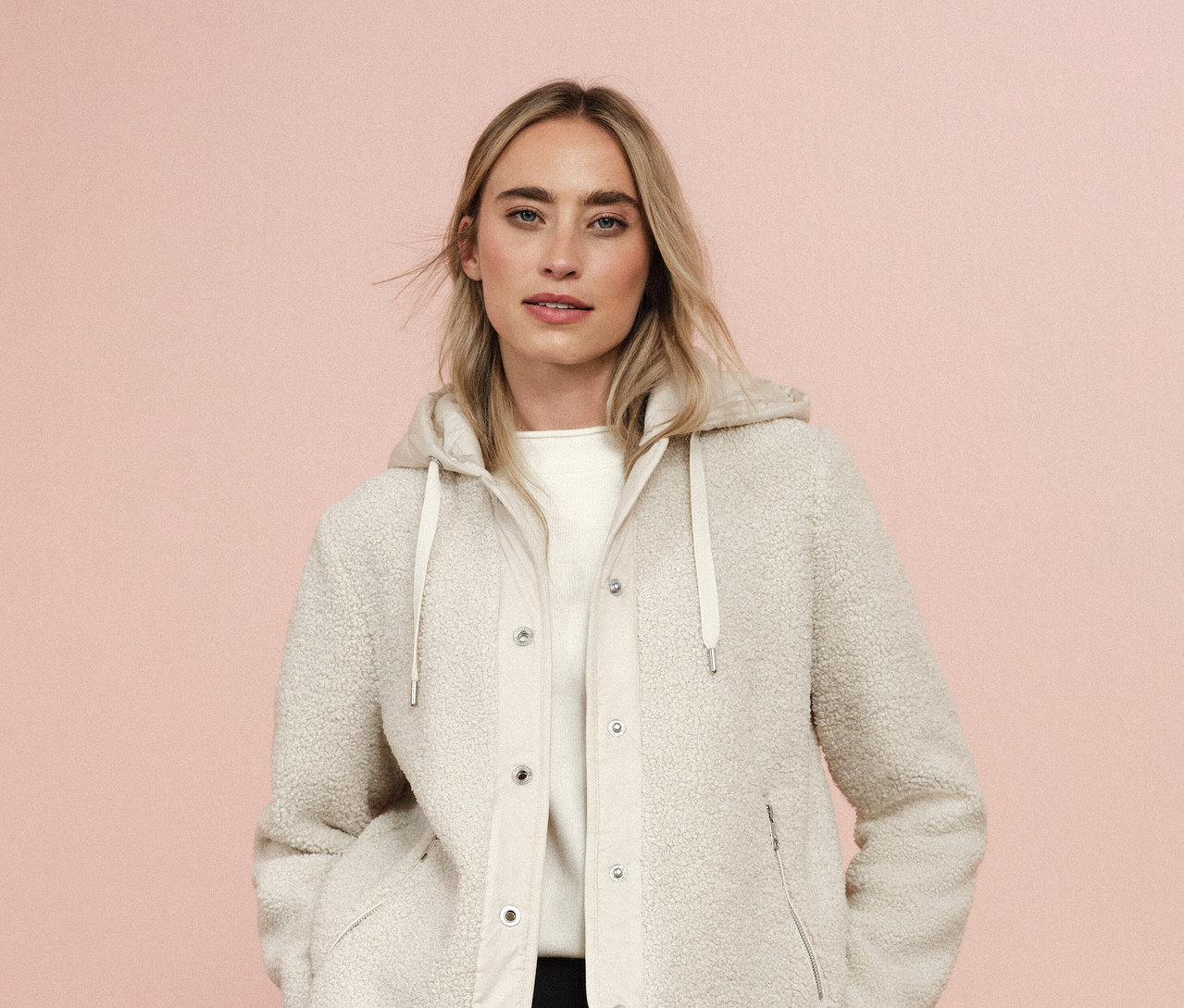 Frau trägt eine Street One Teddyjacke mit abnehmbarer Kapuze vor einem rosafarbenen Hintergrund.