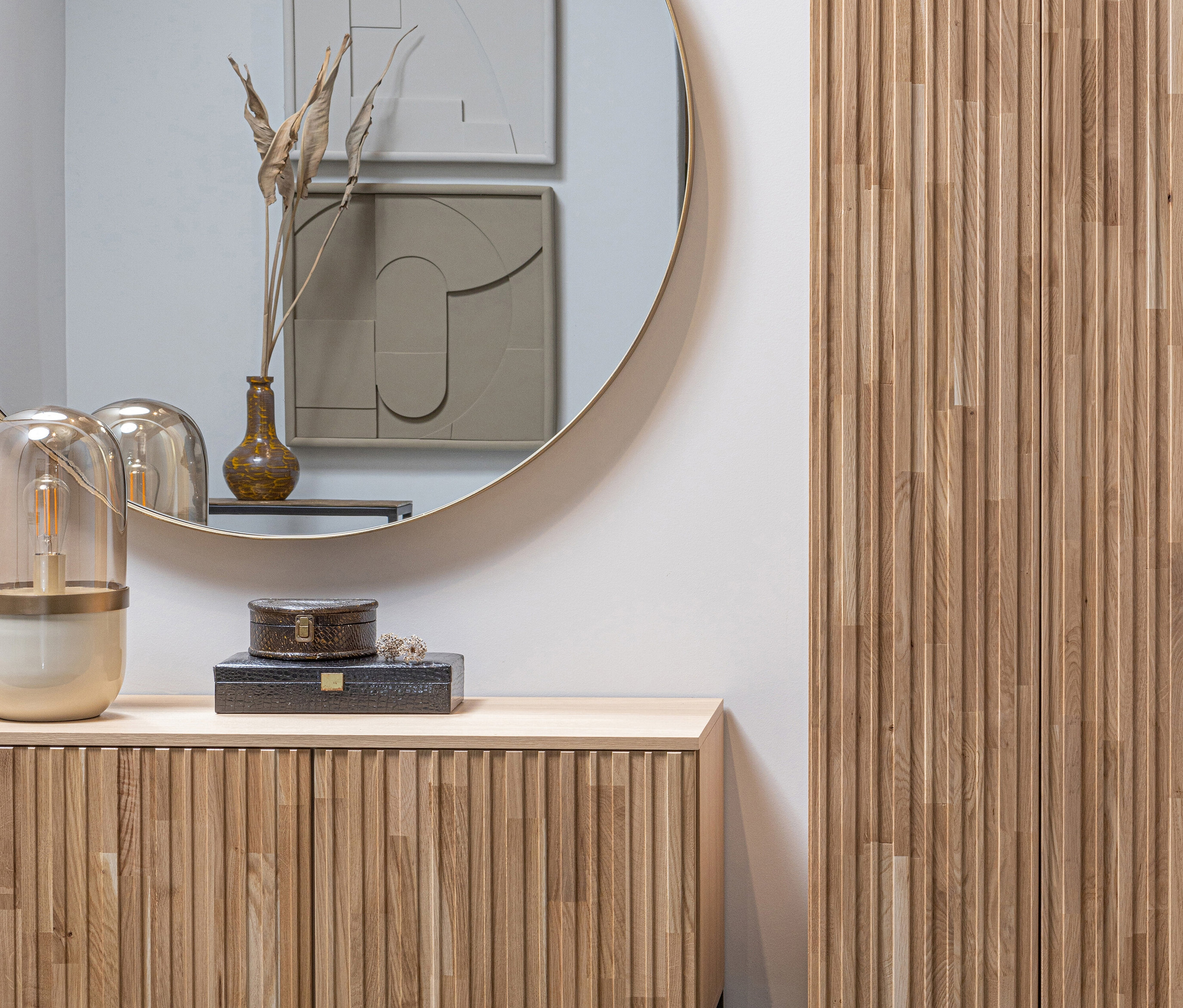 Einrichtung mit einer Kommode aus Holz, auf der dekorative Schachteln und eine Lampe stehen. Im Hintergrund ein runder Spiegel und ein Holzschrank. An der Wand hängt ein goldfarbener, runder Woood Spiegel »Doutzen« mit 115 cm Durchmesser.
