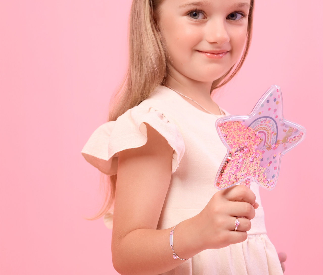 Ein lächelndes Mädchen mit blonden Haaren hält einen sternförmigen Zauberstab. Sie trägt ein Amor 925 Silber Kinder-Armband mit Herz-Anhänger und Plättchen am linken Handgelenk und einen pinken Ring am linken Ringfinger.