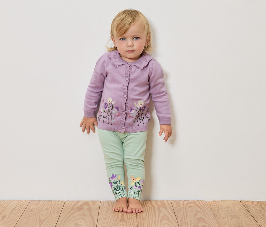 Ein blondes Kleinkind steht barfuß vor einer weißen Wand, trägt eine lila Strickjacke mit Blumenmuster und grüne Leggings mit Blumen am Bein.