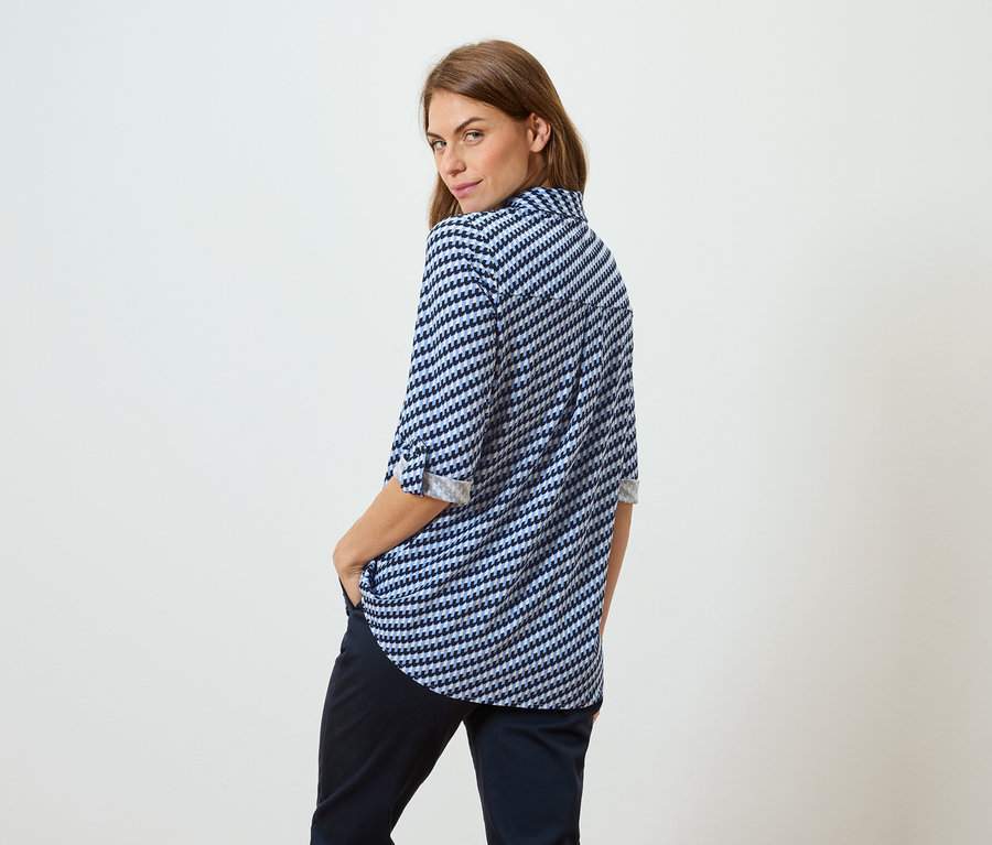 Frau mit braunen Haaren, die eine blau-weiß gemusterte Bluse und dunkle Hosen trägt, steht mit dem Rücken zum Betrachter und blickt über ihre Schulter.