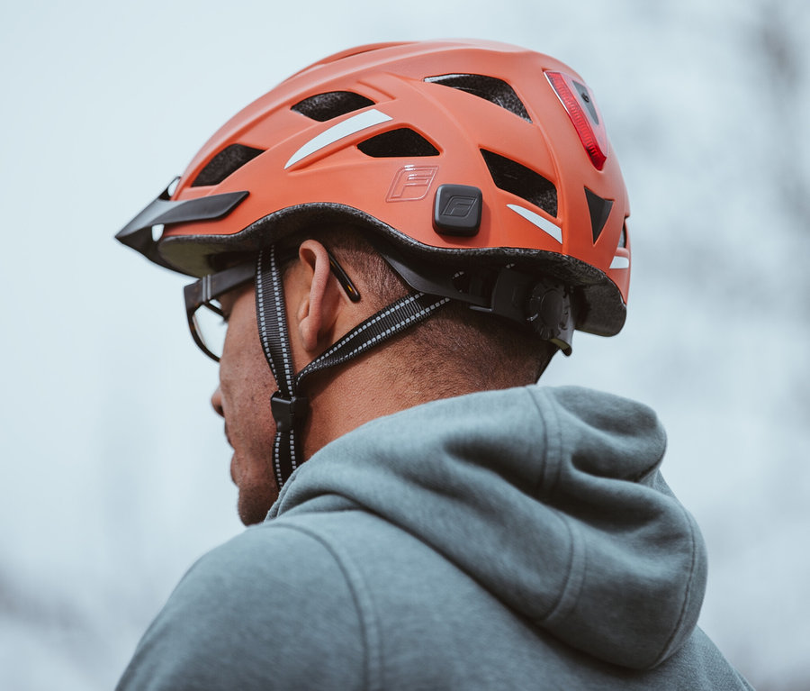 Nahaufnahme eines Mannes mit Brille und grauem Hoodie, der einen orangefarbenen FISCHER Fahrradhelm Urban PLUS Miami trägt.