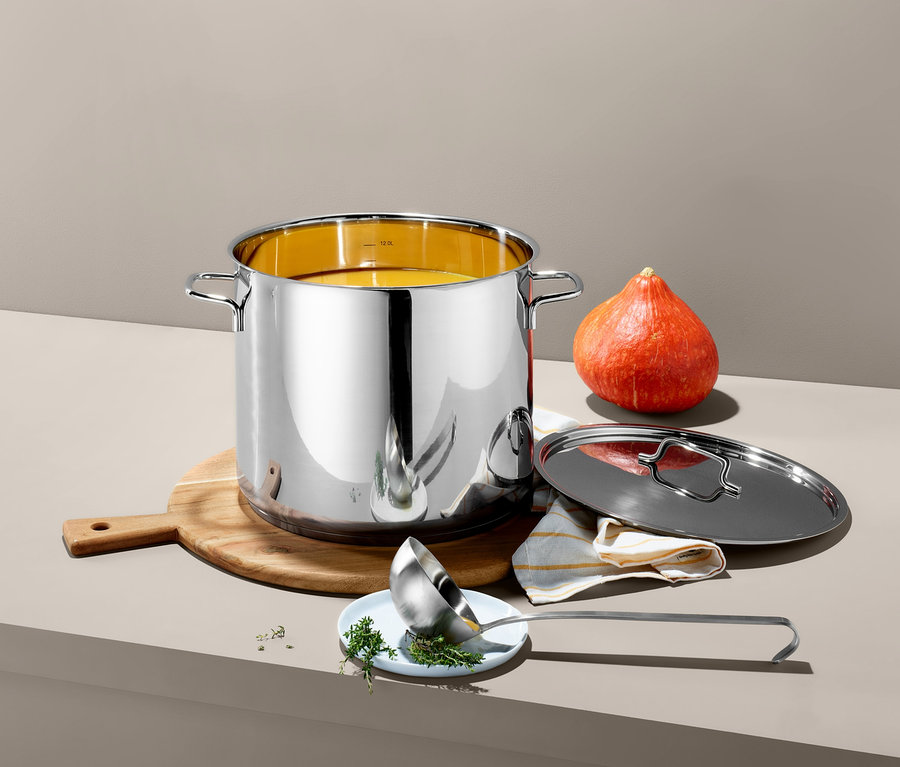 Ein XL-Edelstahl-Topf steht auf einem Holzbrett neben einem Kürbis. Ein Deckel und eine Schöpfkelle liegen neben dem Topf.