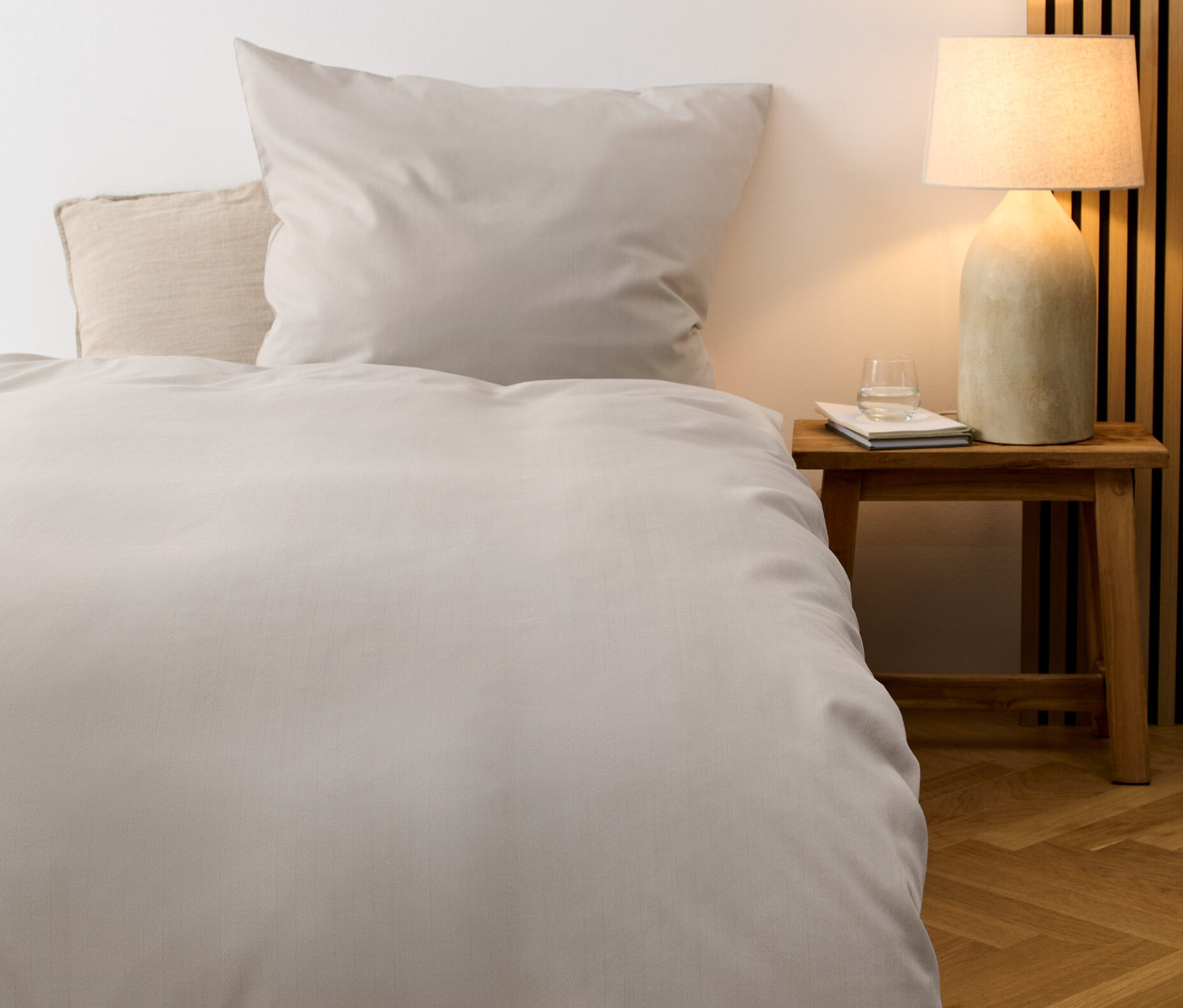 Ein hellgraues Edelflanell-Bettwäsche-Set in Normalgröße liegt auf einem Bett. Ein Nachttisch mit Lampe und Wasserglas steht neben dem Bett.