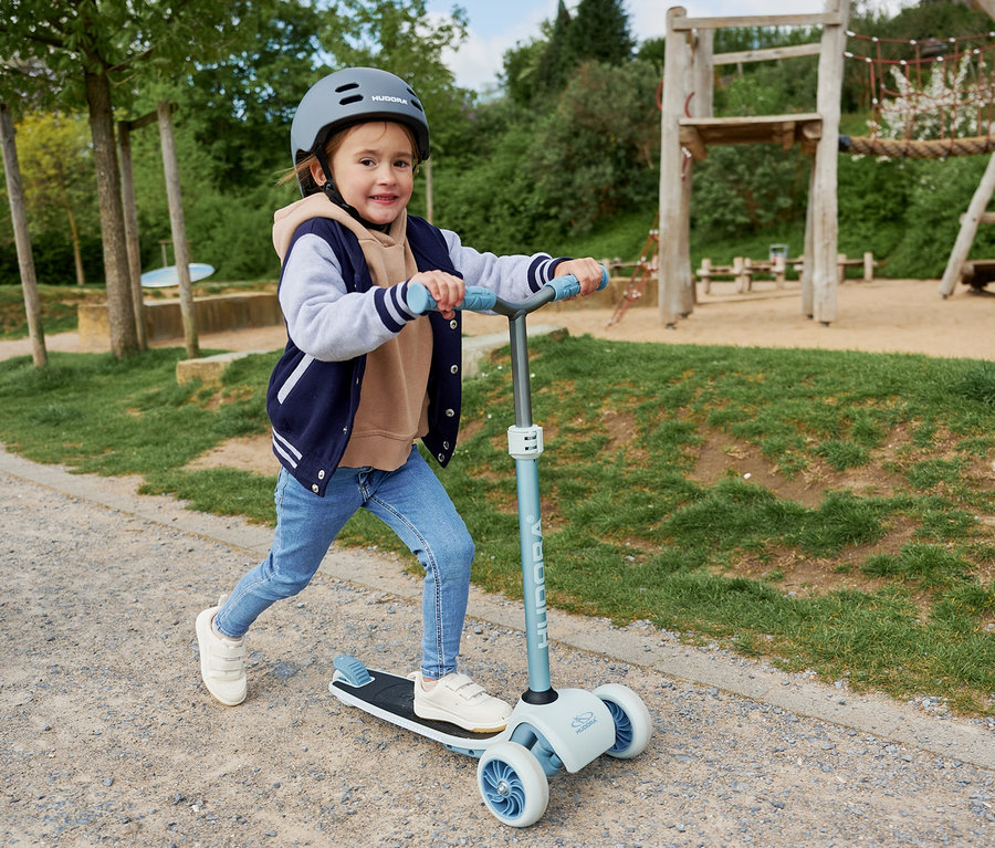 Mädchen fährt mit blauem HUDORA Tri-Scooter auf einem Weg im Park.