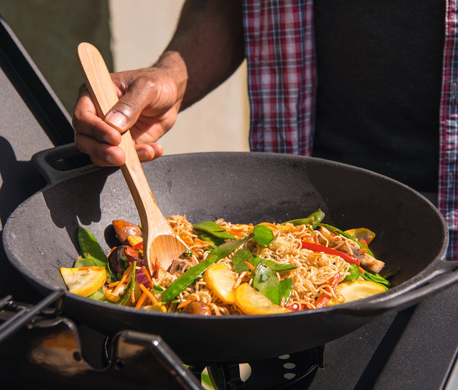 Mann rührt mit einem Holzlöffel Nudeln mit Gemüse in einem Landmann Grillwok.