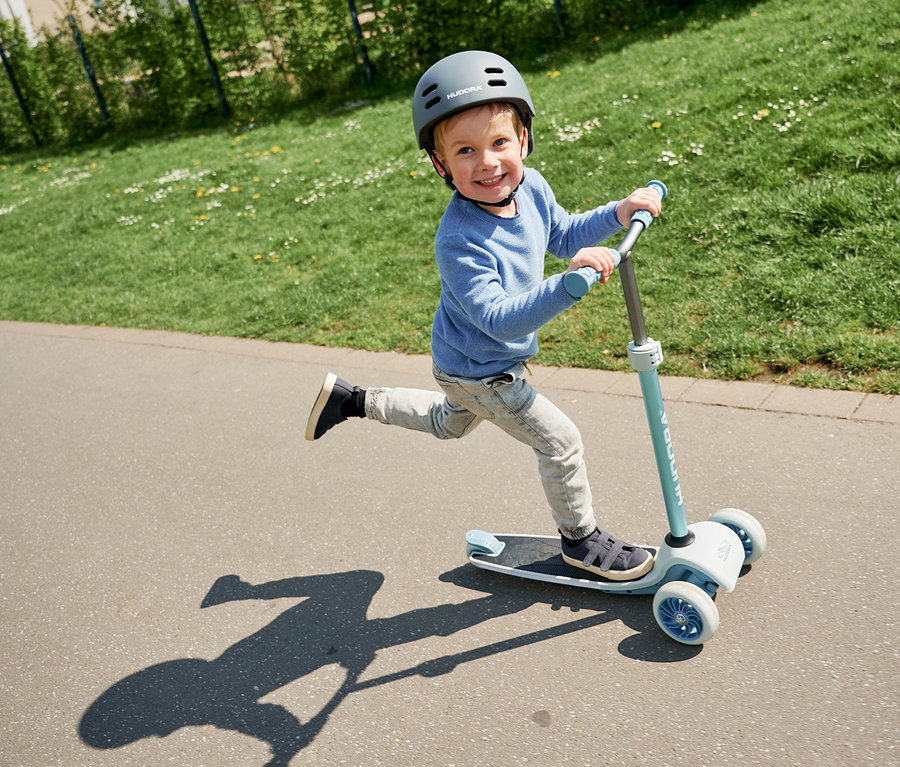 Ein Junge fährt mit einem blauen HUDORA Tri-Scooter und Helm auf dem Kopf auf dem Gehweg.