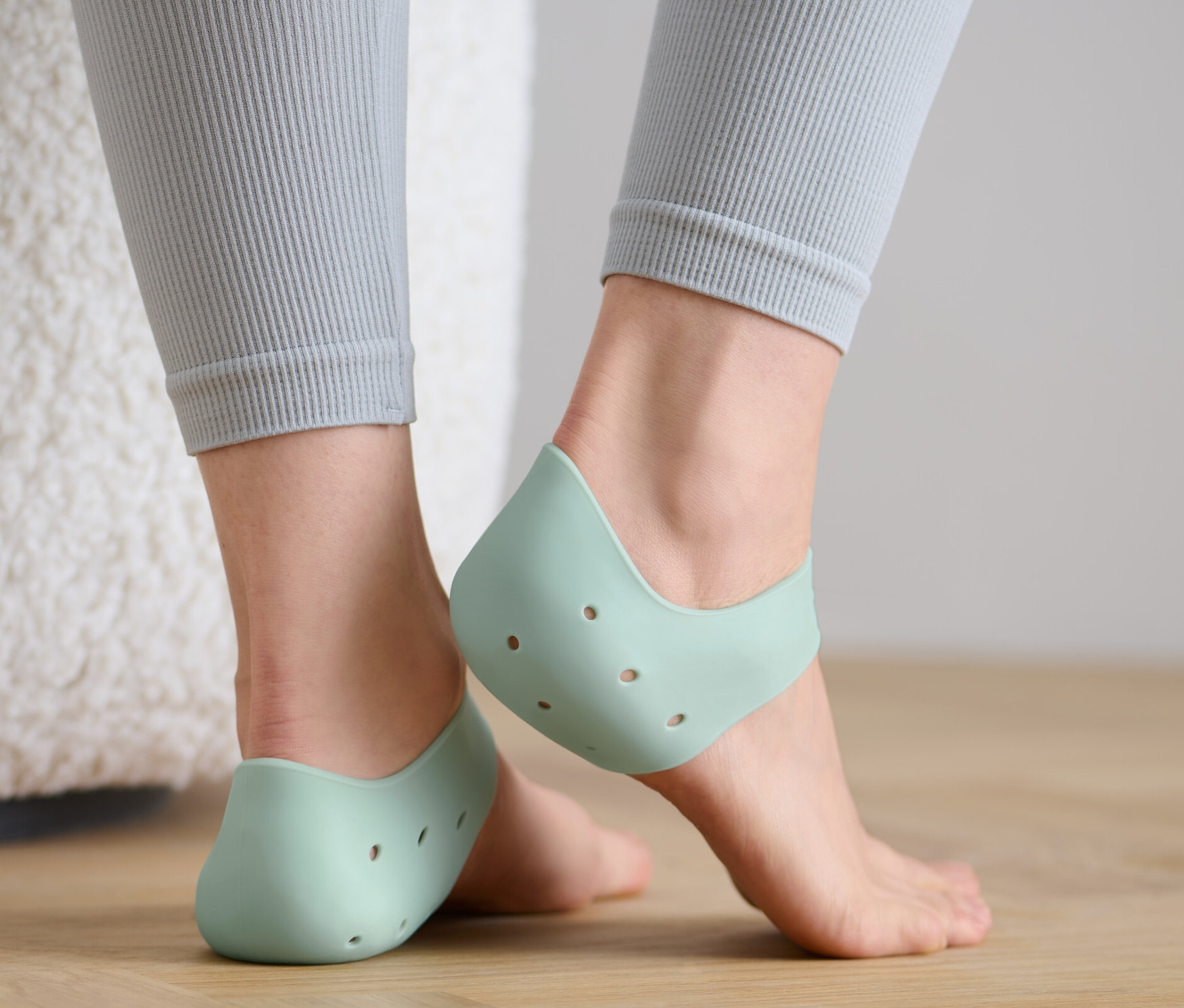 Frauenfüße in grauen Leggings stehen auf einem Holzboden und tragen 2 Fersenschutzhauben.