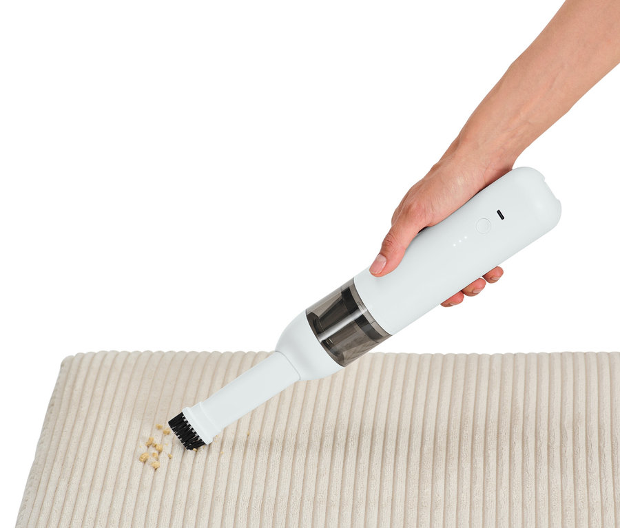Eine Hand verwendet einen Akku-Handstaubsauger, um Krümel von einem cremefarbenen Stoff zu entfernen.