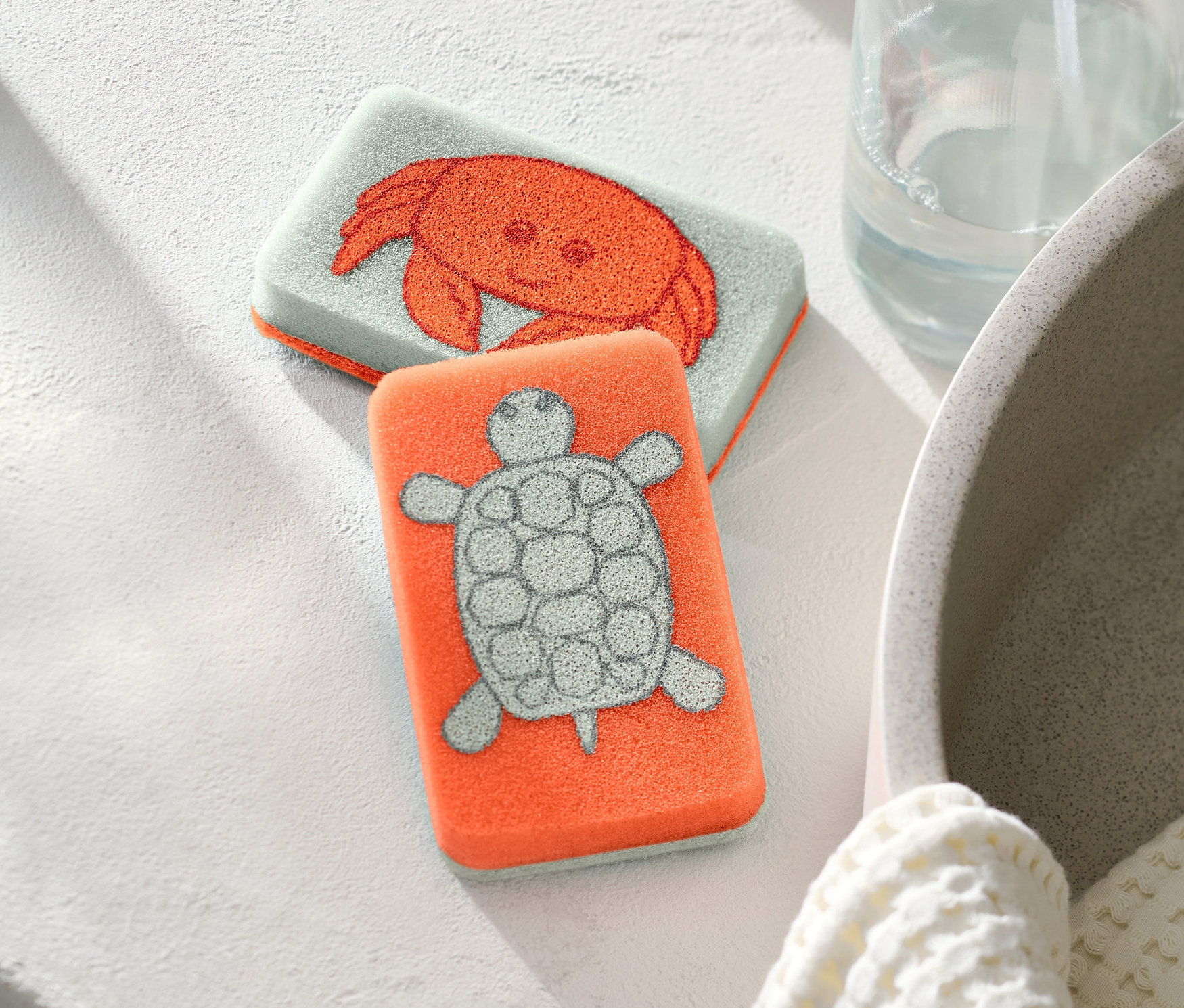 Zwei Reinigungsschwämme, einer mit einem Krabbenmotiv und einer mit einem Schildkrötenmotiv, liegen nebeneinander.