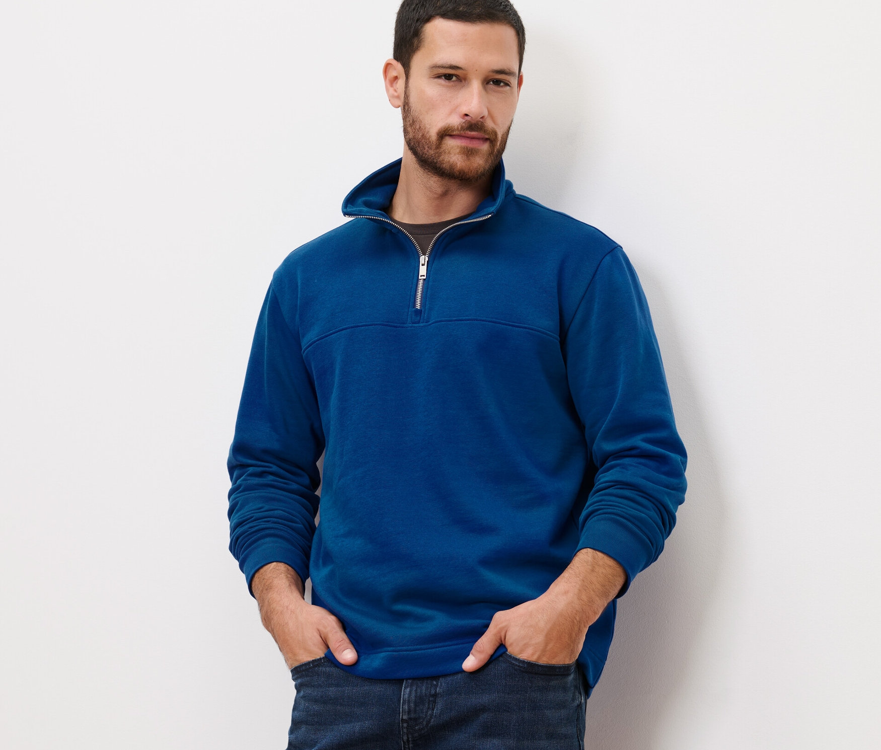 Mann mit den Händen in den Taschen trägt einen blauen Sweat-Troyer und Jeans.