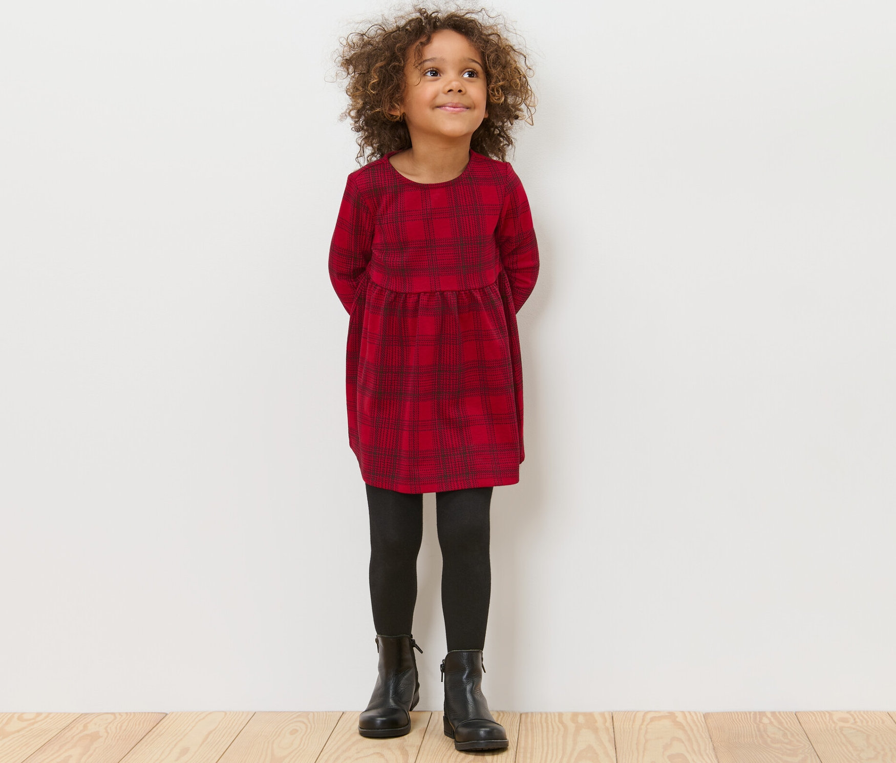 Ein Mädchen steht mit den Händen hinter dem Rücken und trägt ein Kinder-Jersey-Kleid, eine schwarze Leggings und schwarze Schuhe.