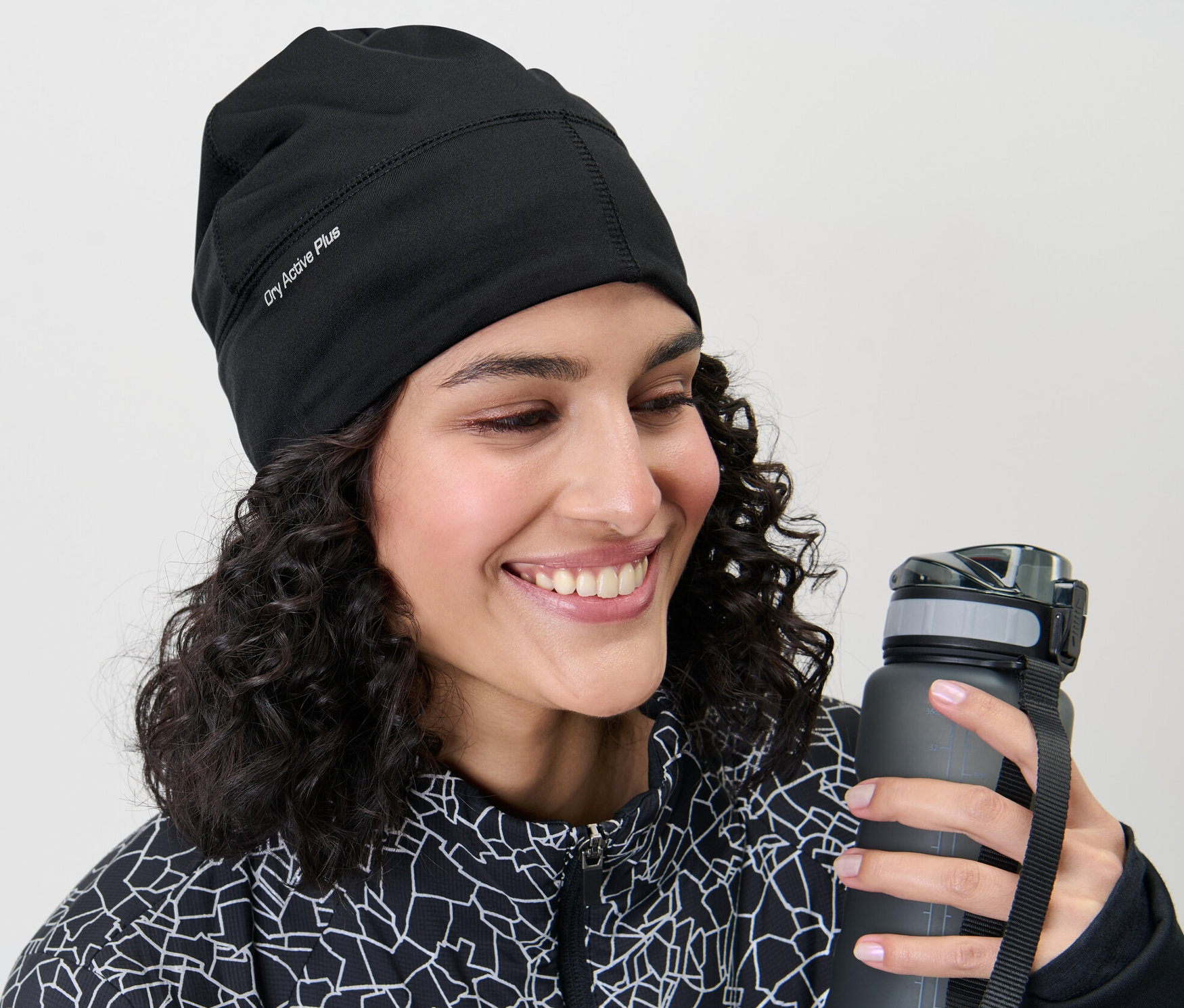 Lächelnde Frau mit lockigem Haar hält eine graue Wasserflasche und trägt eine schwarze Thermo-Funktionsmütze.
