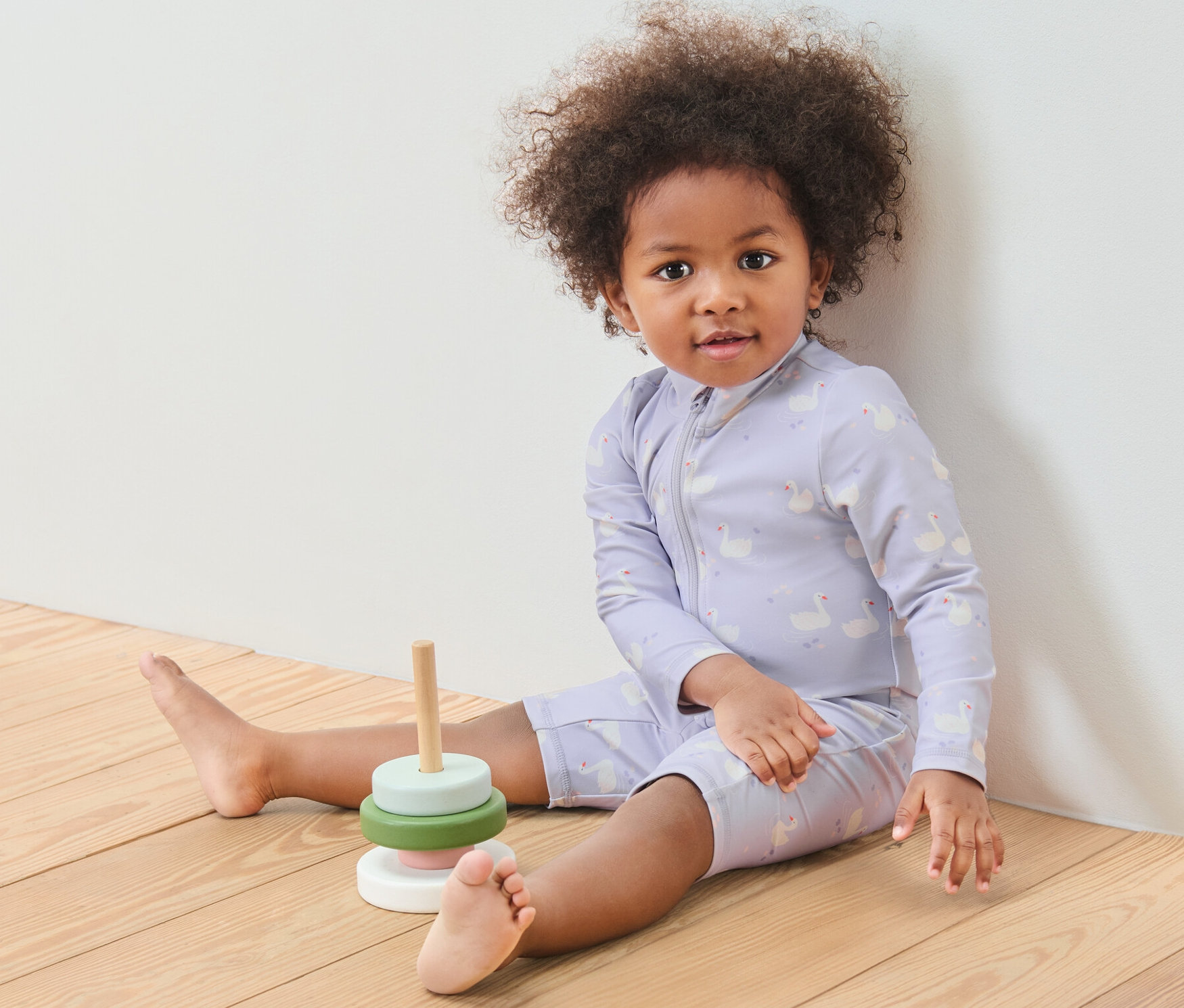 Ein Kind sitzt auf einem Holzboden neben einem Holzspielzeug. Es trägt einen Baby-Einteiler mit UV-Schutzfaktor 80, Schwan.