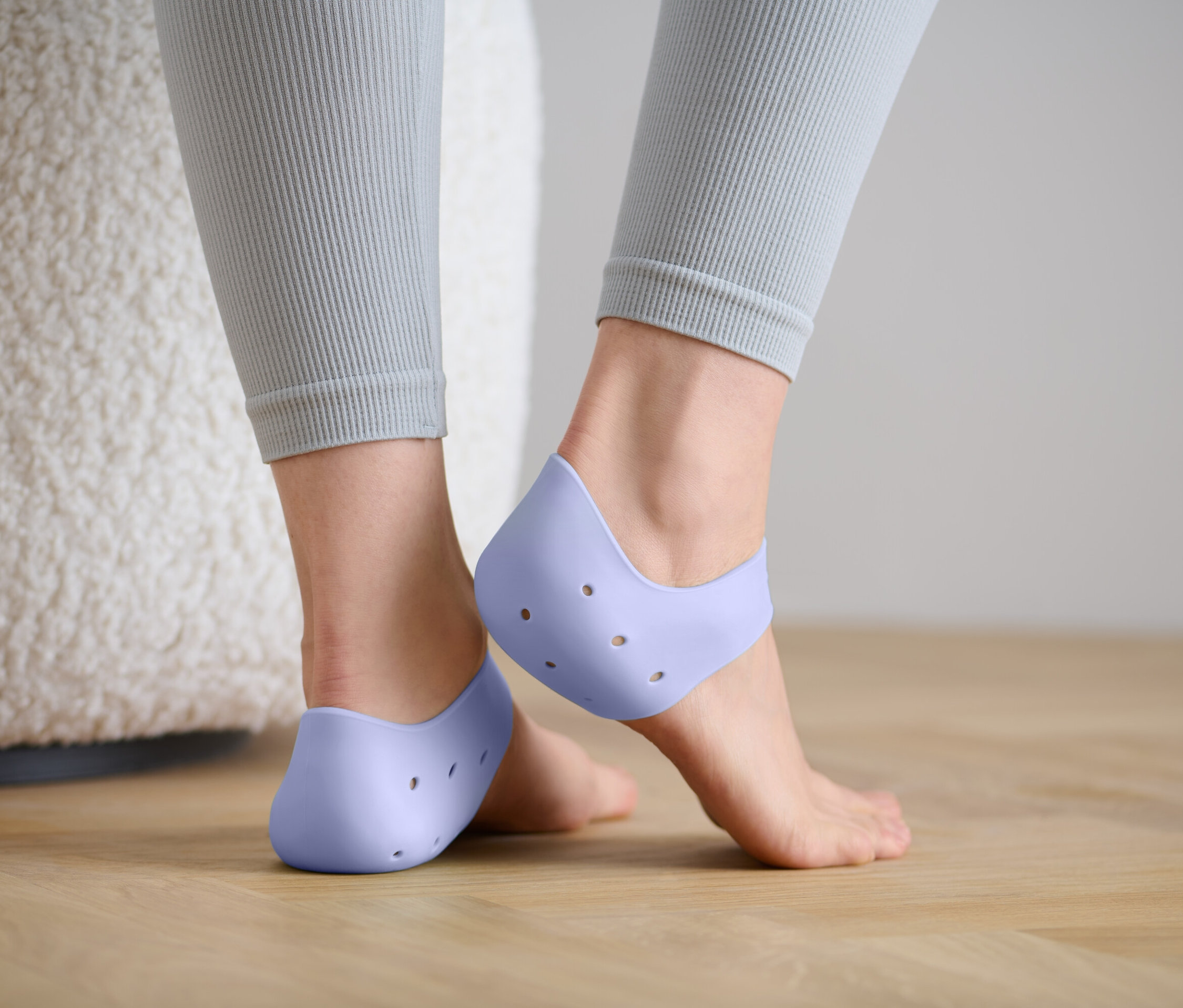 Rückansicht einer Frau in grauen Leggings, die hellviolette Silikon-Fersenschoner mit Löchern trägt.