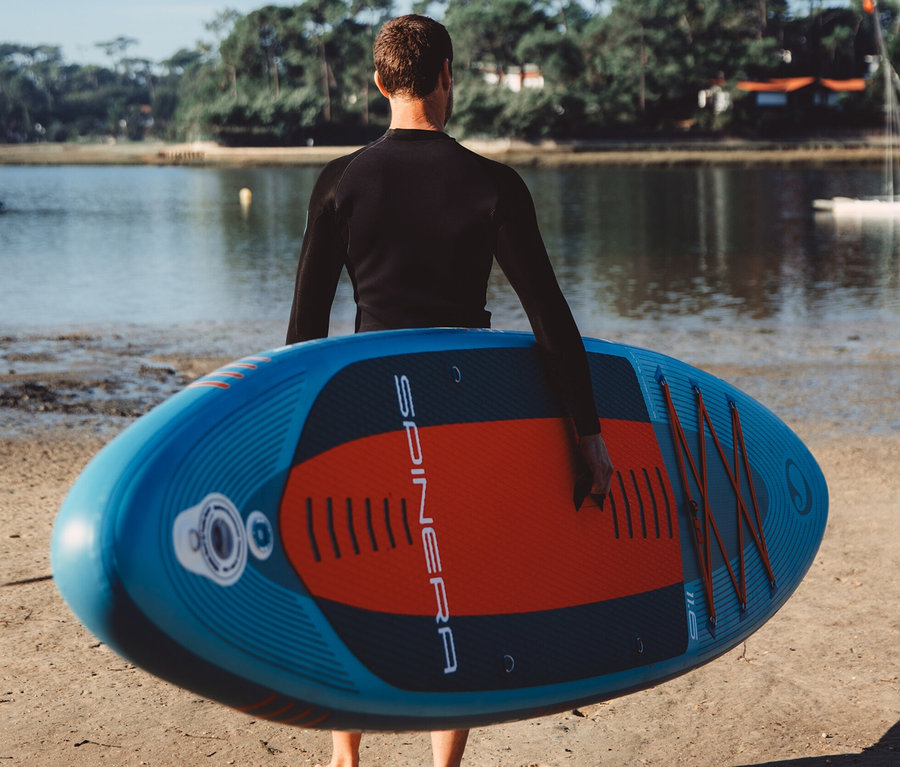 Mann im Neoprenanzug trägt ein Spinera SUP Board Lets Paddle 11' 6" am Strand.