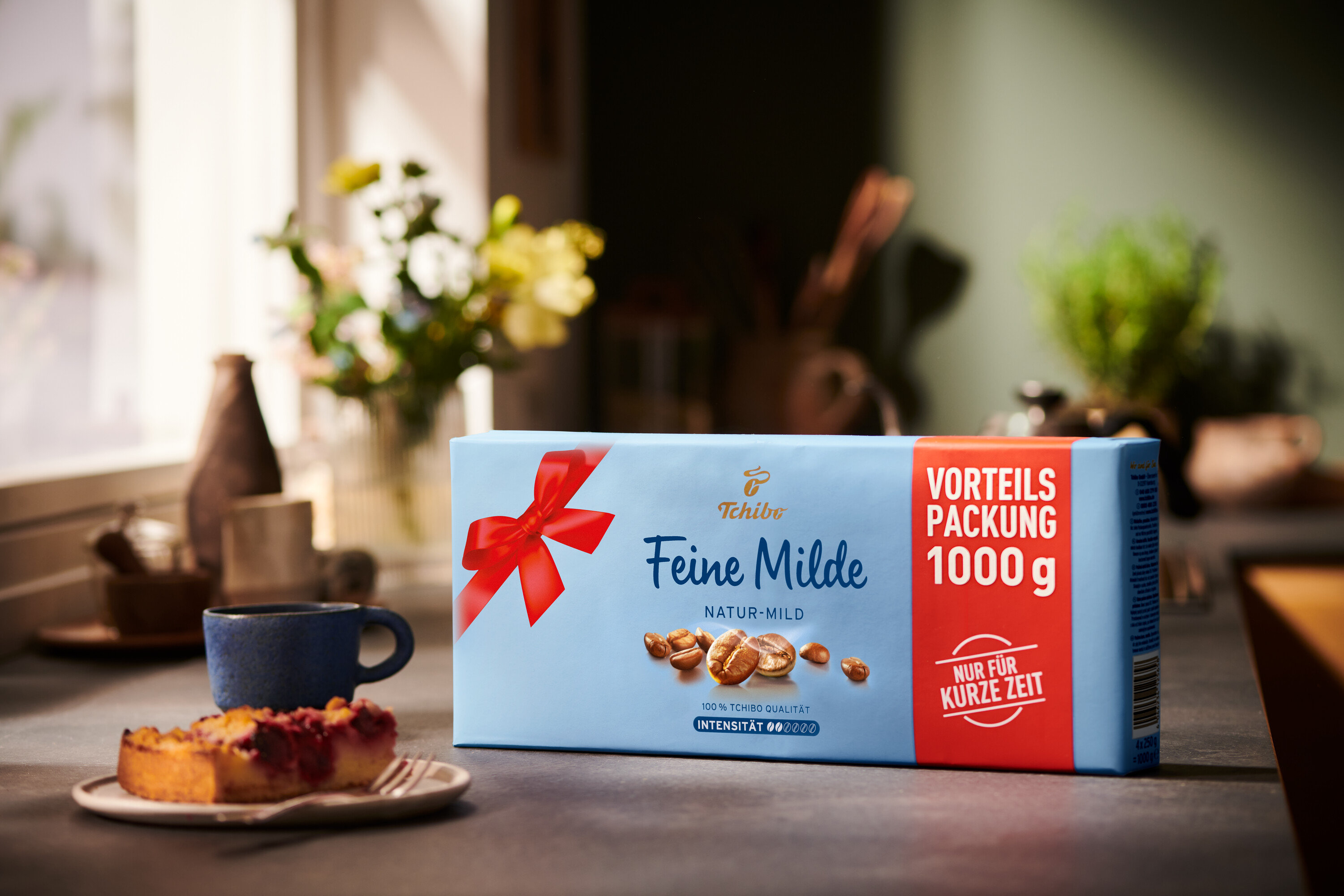 Eine Packung Feine Milde Kaffee steht neben einer Tasse und einem Stück Kuchen auf einem Tisch.