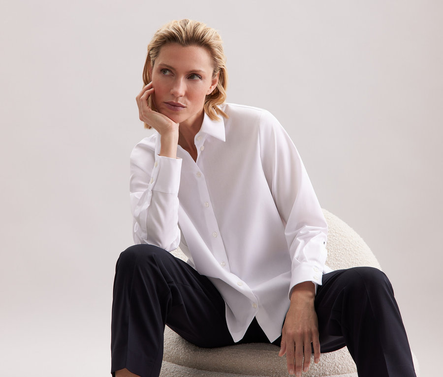 Frau sitzt auf einem Stuhl, trägt ein weißes Hemd und eine schwarze Hose.