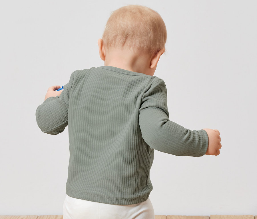Kleinkind von hinten trägt ein hellgrünes geripptes Langarmshirt und eine weisse Hose.