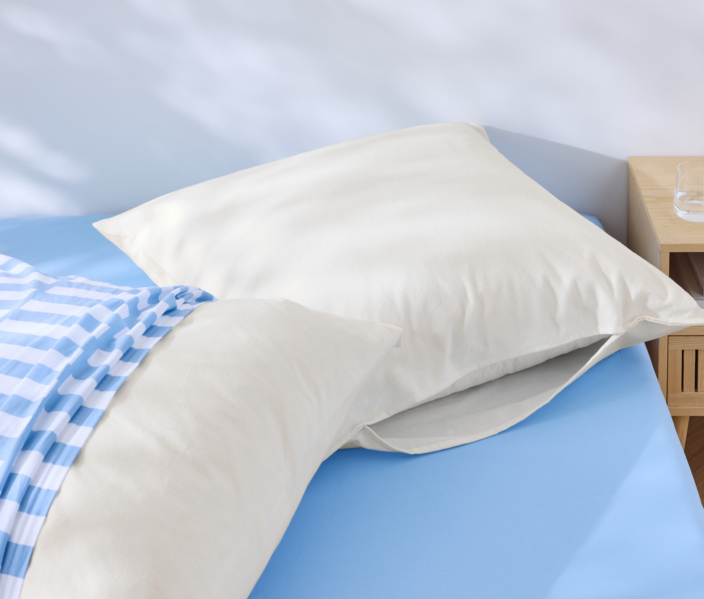 Zwei weiße Kopfkissen liegen auf einem blauen Bett, eines davon mit einem blau-weiß gestreiften Bezug. Neben dem Bett steht ein Nachttisch mit Wasser und einem Telefon.