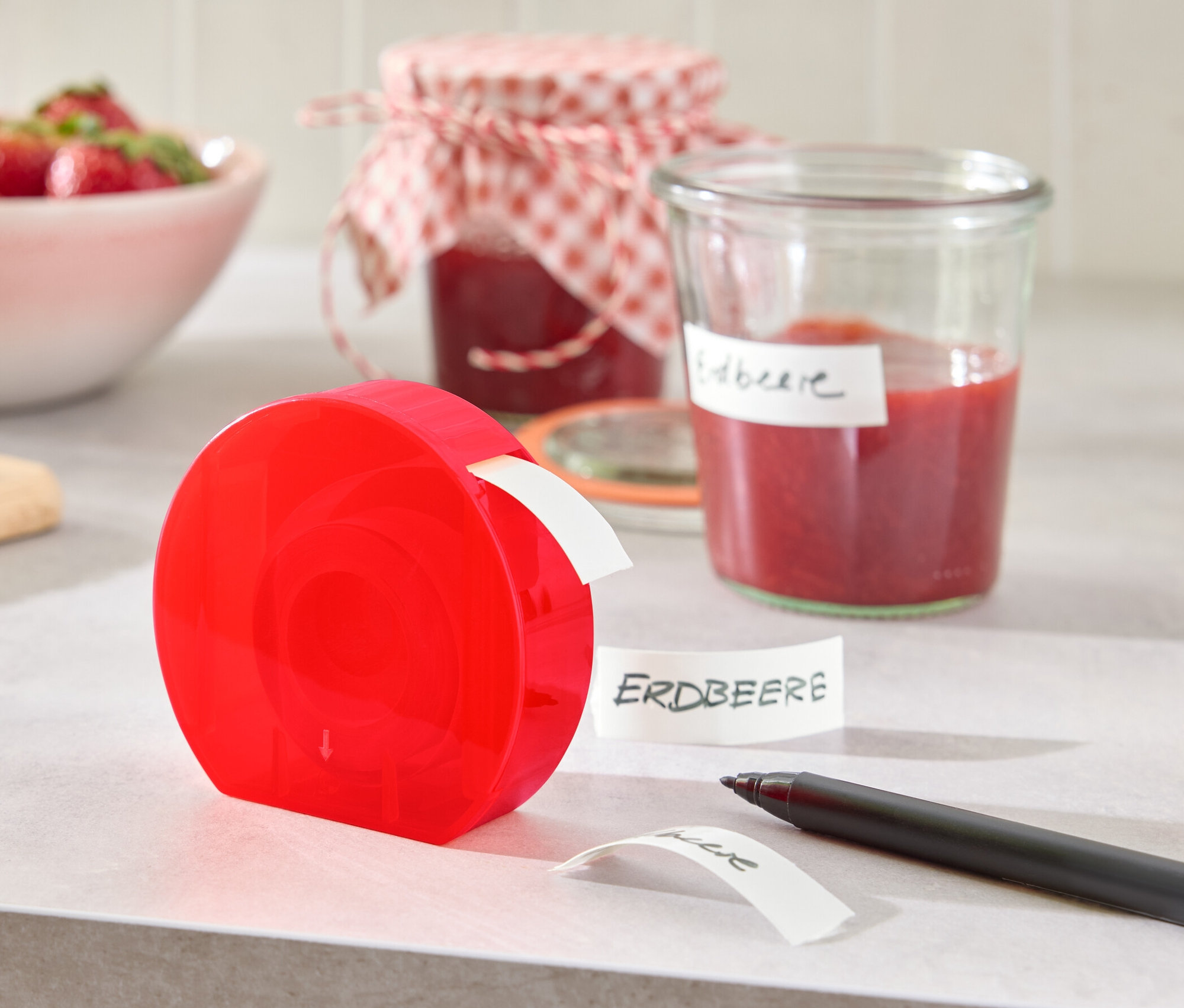 Ein roter Etikettenspender steht auf einem Tisch neben einem Glas Marmelade und einer Schüssel Erdbeeren.