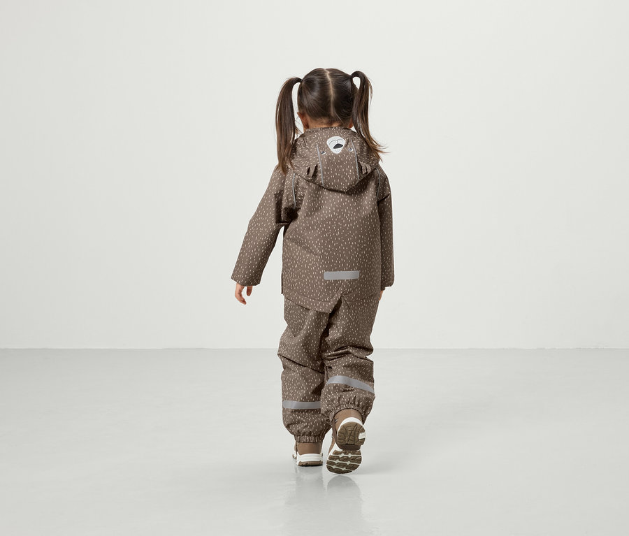 Mädchen mit Zöpfen geht in brauner Kapuzenjacke und Hose über den Boden.