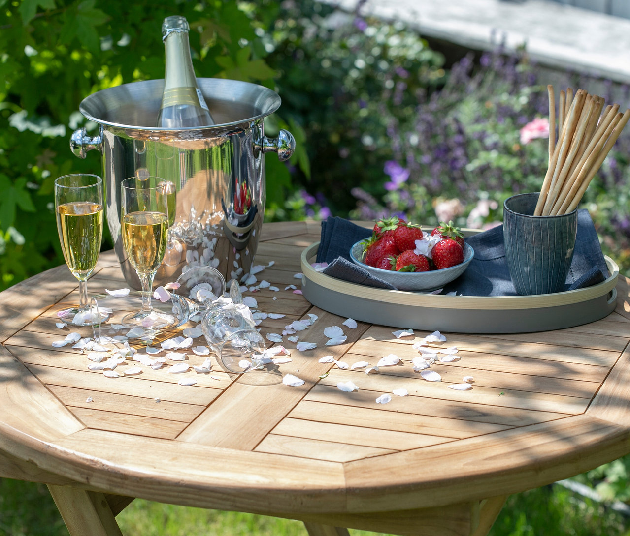 Auf einem Holztisch stehen ein Sektkübel mit einer Flasche, zwei gefüllte Sektgläser, umgefallene Gläser, Erdbeeren, Grissini und Blütenblätter.