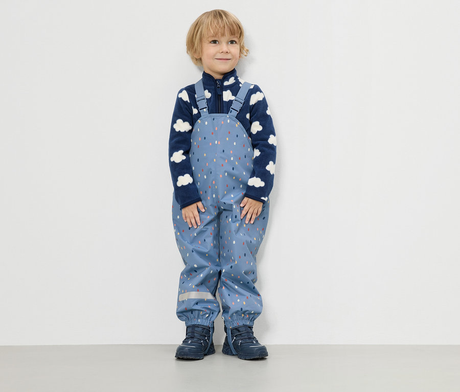 Ein kleiner Junge steht vor einer weißen Wand und trägt eine blaue Fleecejacke mit Wolkenmuster und eine blaue Regenhose mit bunten Tropfen.
