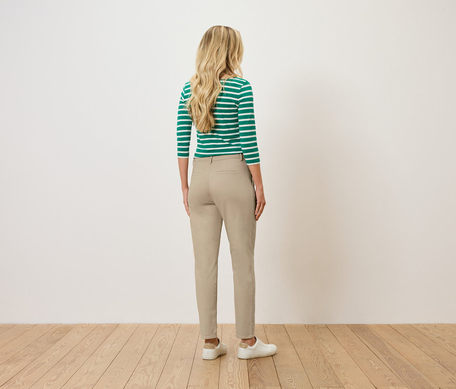 Eine Frau mit blonden, gewellten Haaren steht mit dem Rücken zum Betrachter und trägt ein grün-weiß gestreiftes Oberteil, eine beige Hose und weiße Turnschuhe.