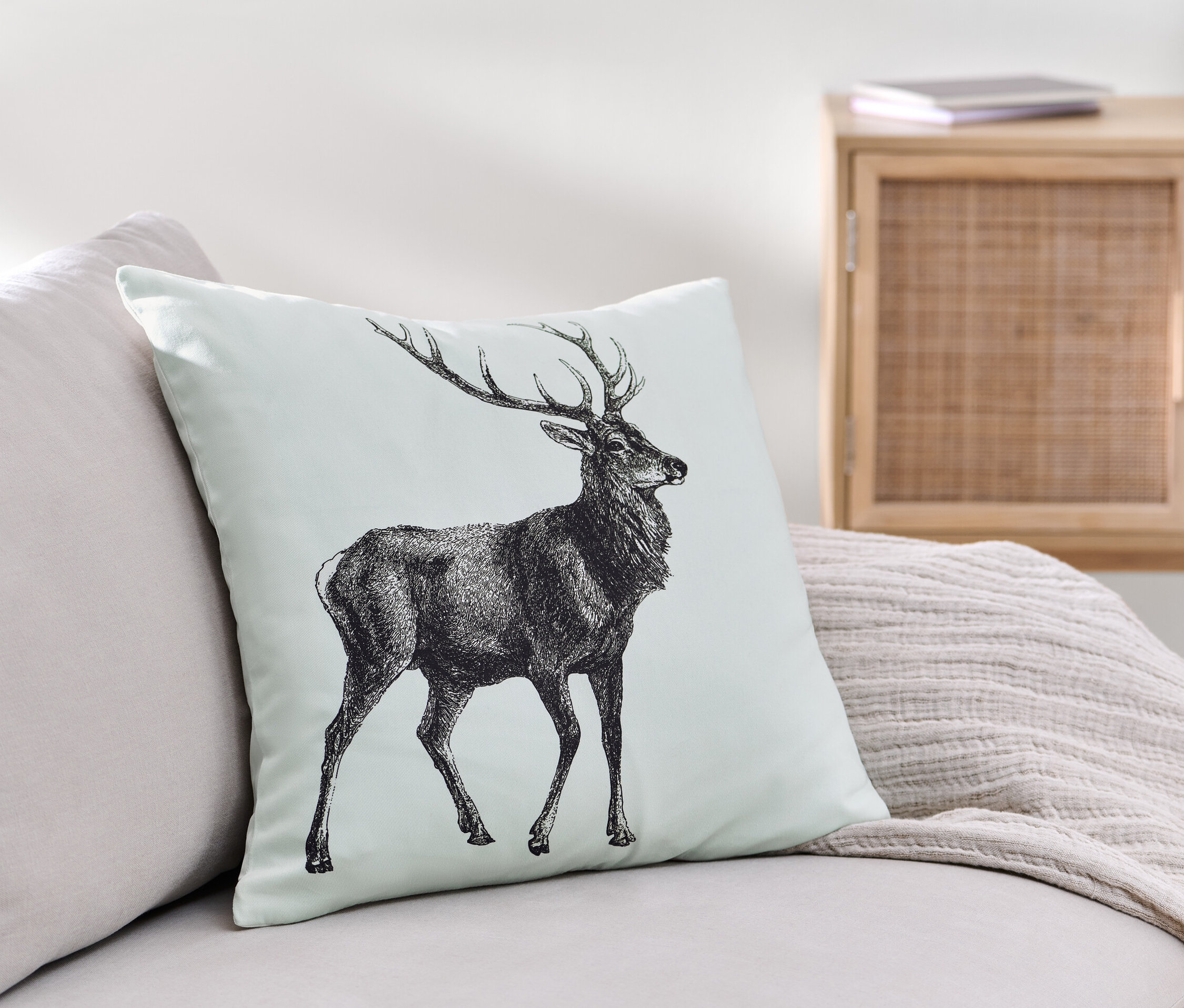 Ein Dekokissen-Bezug mit einem Hirschmotiv liegt auf einem Sofa.