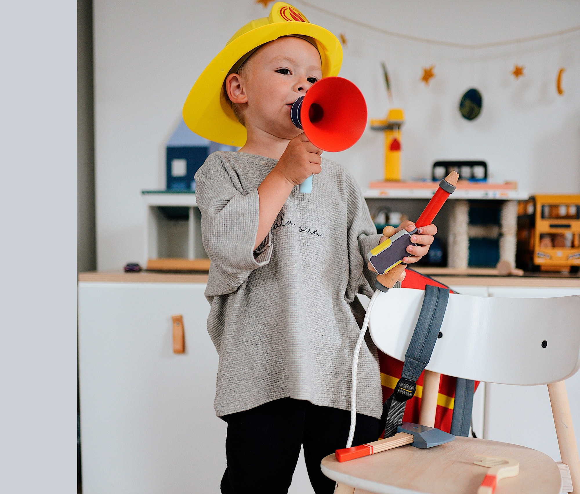 Ein Junge mit Feuerwehrhelm und Megafon. Auf einem Stuhl liegt eine Feuerwehraxt und dahinter ist ein small foot Feuerwehrrucksack zu sehen.
