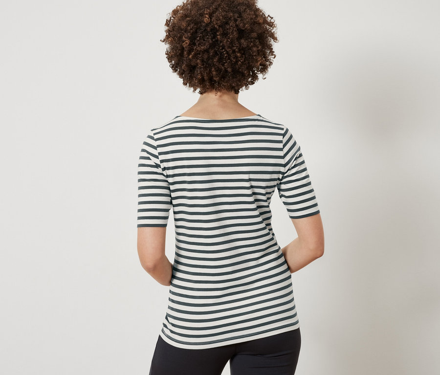 Rückansicht einer Frau mit braun gelocktem Haar, die ein grün gestreiftes Halbarmshirt und eine schwarze Hose trägt.