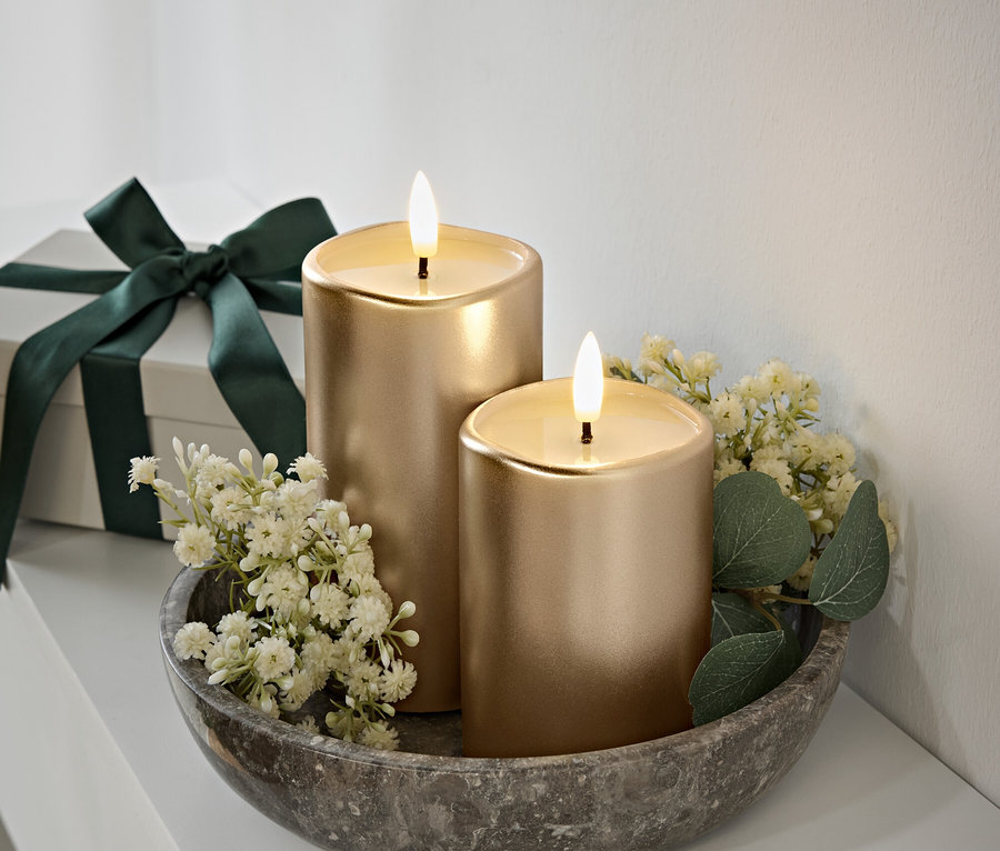 Zwei goldene Kerzen mit Flammen in einer grauen Steinschale, weiße Blüten und grüne Blätter. Geschenkbox mit dunkelgrünem Band im Hintergrund.