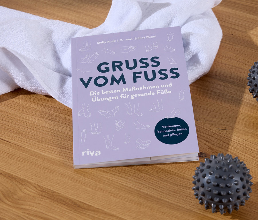 Das Buch Gruss Vom Fuss neben einem weißen Handtuch und zwei Massagebällen auf einem Holzboden.