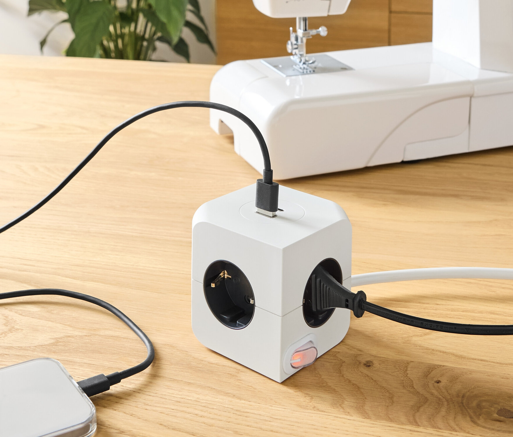 Auf einem Holztisch steht ein Steckdosenwürfel mit USB, an den ein Telefon und eine Nähmaschine angeschlossen sind.