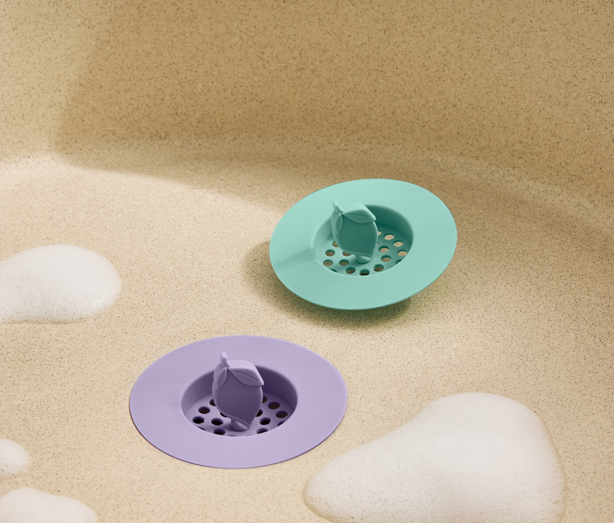 Draufsicht auf einen grünen und einen lilafarbenen Waschbeckenstöpsel mit Schaum in einem beigen Waschbecken.