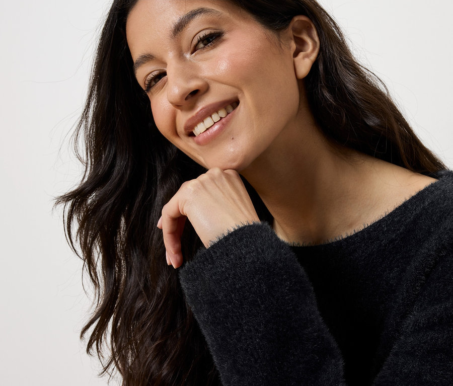 Lächelnde Frau mit langen, gewellten braunen Haaren, die ihr Kinn auf ihre Hand stützt und einen schwarzen Pullover trägt.